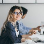 Attraktive lachende Frau mit Brille und blonden Haaren posiert mit einer Tasse Kaffee an ihrem Arbeitsplatz. Das Foto steht repräsentativ für eine Dolmetscher.