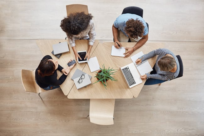 Sicht von oben. Vier Mitarbeitende, darunter drei Frauen und ein Mann halten ein Meeting. Auf dem Tisch liegen Schreibblöcke und mobile Geräte wie Tablet und Laptop.